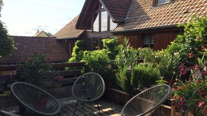 A wooden deck with wicker chairs, surrounded by lush greenery and a house with a sloped roof.