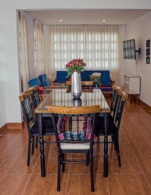Ein Essbereich mit einem runden Tisch, sechs Stühlen, einer Vase mit Blumen und einem blauen Sofa im Hintergrund.