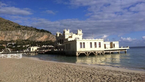 Un edificio costero con balcón que da a la playa, un muelle que se adentra en el agua y montañas al fondo.