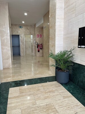 Un hall d’entrée avec un sol en marbre, une plante en pot, des extincteurs et des panneaux indiquant les sorties de secours.