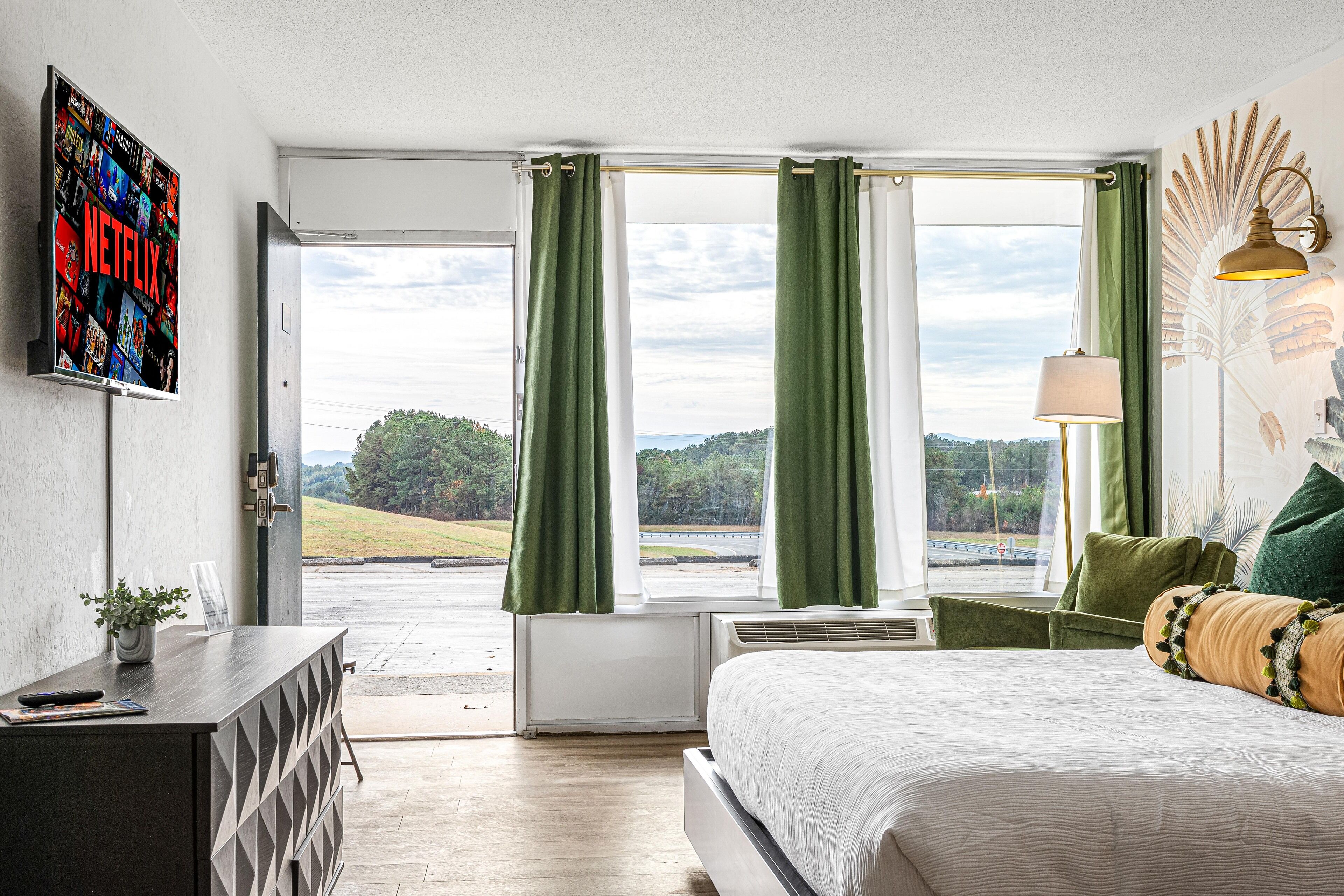 A hotel room with a bed, a desk with a plant, a wall-mounted TV displaying Netflix, and a view of a road and greenery through large windows.