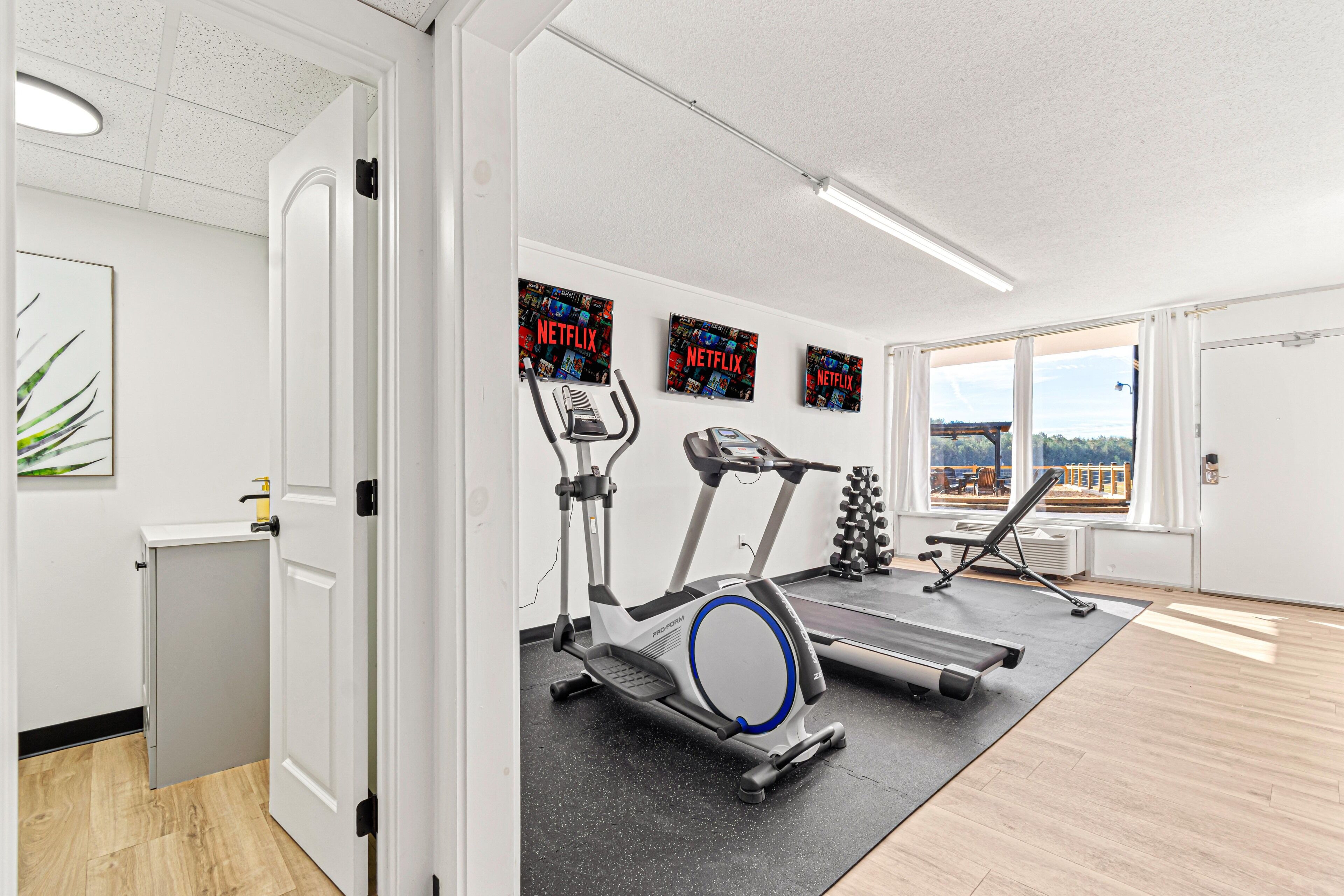 A modern gym with exercise equipment, a treadmill, and a stationary bike. There are large windows with a view of the outdoors.
