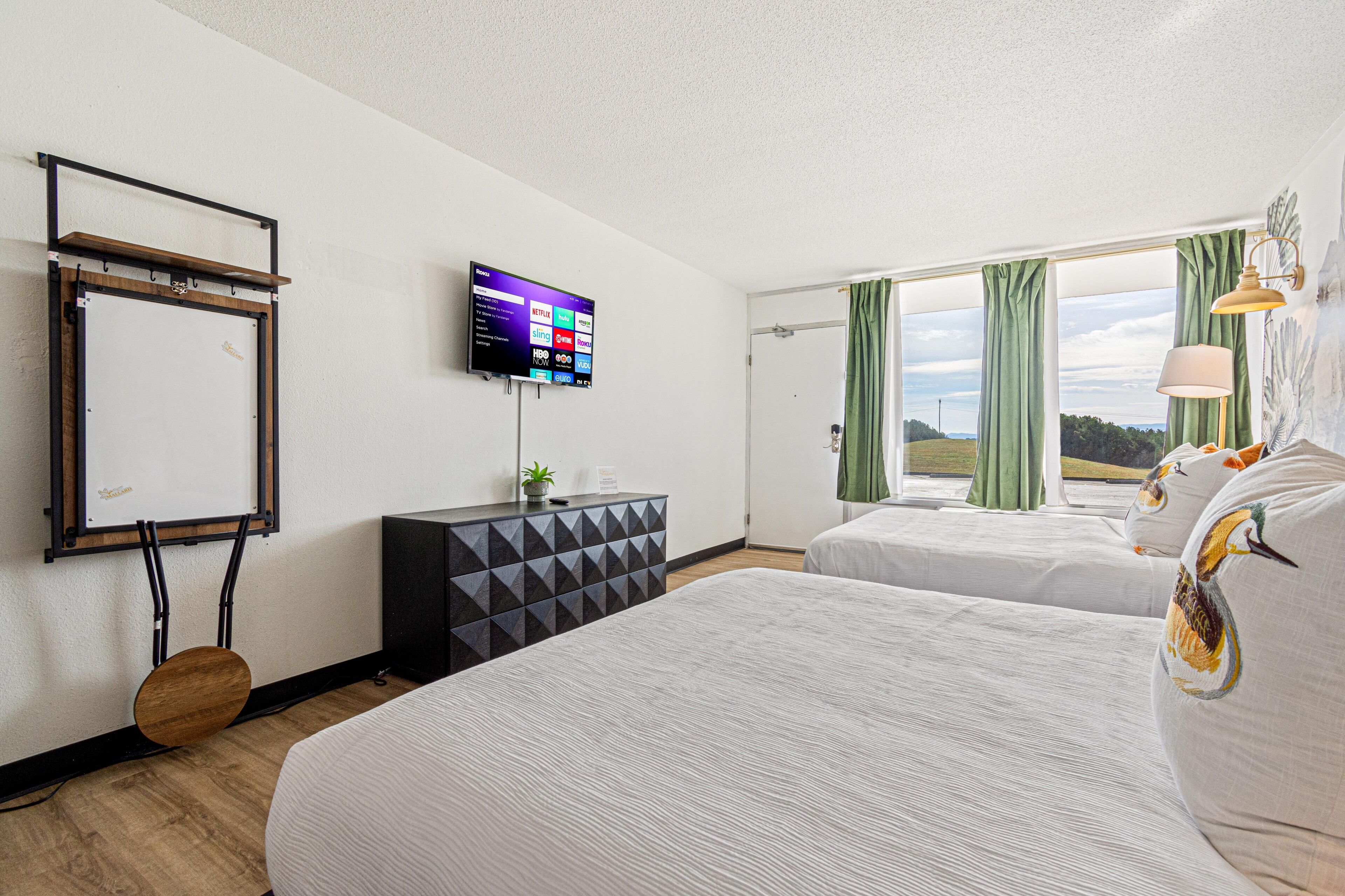 A hotel room with two beds, a TV mounted on the wall, a desk with a plant, and a window with curtains.