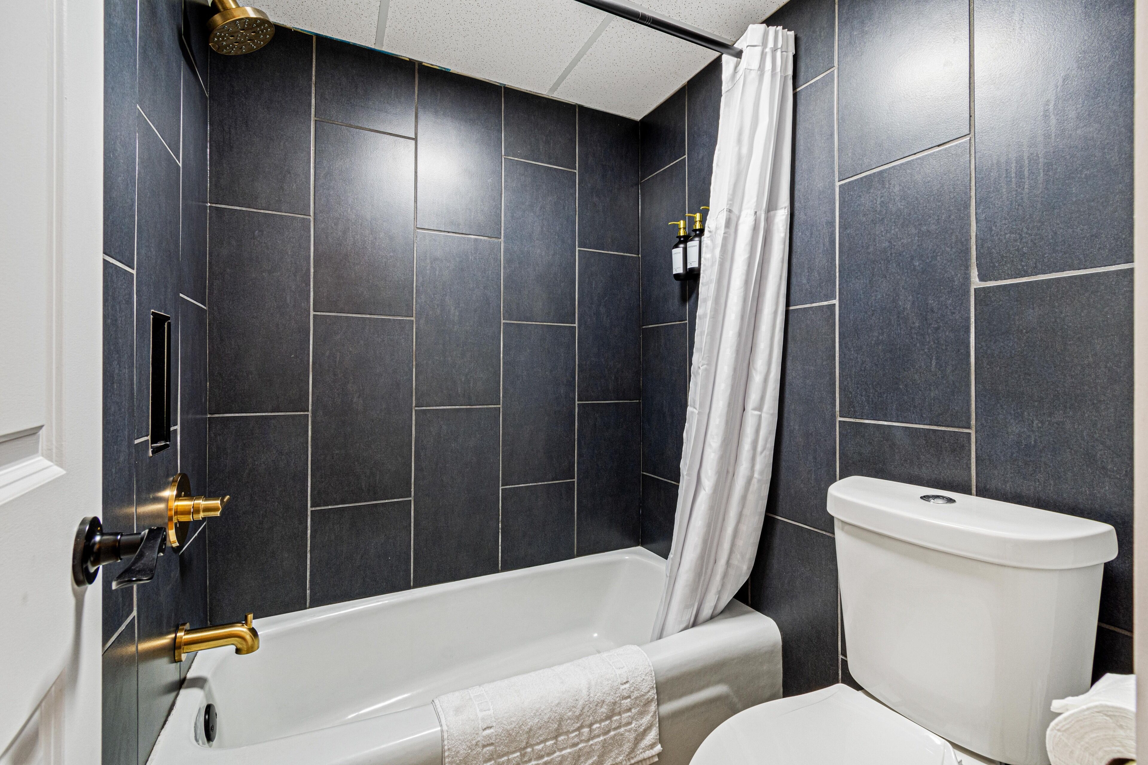 A modern bathroom with dark tiles, a white bathtub, a white toilet, and a shower curtain.
