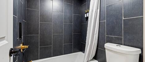 A modern bathroom with dark tiles, a white bathtub, a white toilet, and a shower curtain.