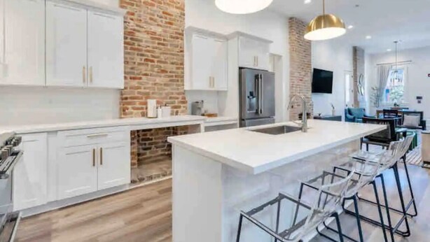 Une cuisine moderne avec des meubles blancs, un mur d’accent en briques, des appareils en acier inoxydable et un îlot central doté de tabourets de bar.