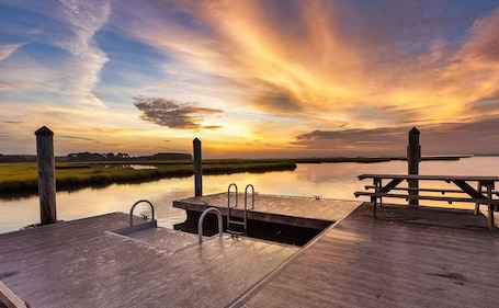 A wooden dock with a bench and a small pool, overlooking a calm body of water during sunset.
