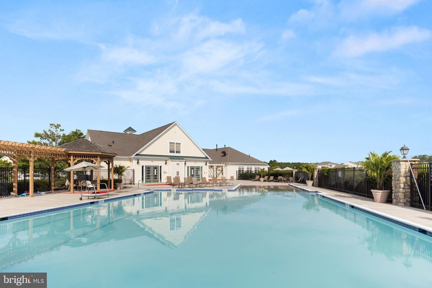 A swimming pool with lounge chairs and a gazebo, surrounded by a fence and a building with a gabled roof.