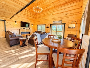 A cozy log cabin interior with a fireplace, a sofa, a dining table, and a television. - Fall Deals/Mobility Friendly/Pets/Games/Secluded (Pigeon Forge)