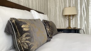 Ein ordentlich bezogenes Bett mit einem dunkel gemusterten Kissen und einem weißen Kissen, eine goldfarbene Lampe auf einem Nachttisch und eine Wand mit vertikaler Holzmaserung.