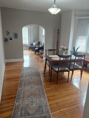 Een eetruimte met een houten tafel en stoelen, een vloerkleed dat naar de eethoek leidt, en een hal met een plafondventilator.