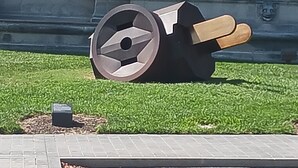 A large, abstract sculpture with a circular base and wooden handles, located in a grassy area with a paved walkway nearby.