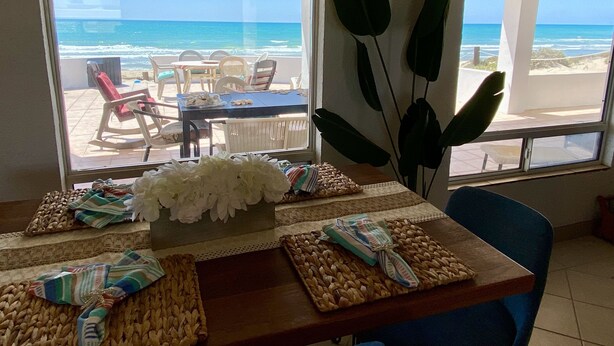A dining area with a view of the beach, a table set with placemats and a centerpiece, and a chair with a striped cushion. - Beach Front Home in beautiful Playa Encanto gated community 3bed/3bath (Playa Encanto)
