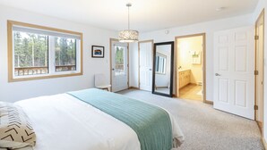 Un dormitorio con una cama grande, un ventanal con vistas a árboles, una puerta que da al baño y una lámpara colgante.