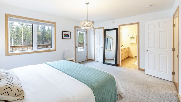 A bedroom with a large bed, a window with a view of trees, a door leading to a bathroom, and a hanging light fixture. - Mountain Haus w/ HOT TUB & large backyard w/firepit. Only 7 mins to downtown (Whitehorse)