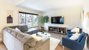 Una sala de estar moderna con un sofá gris, un sillón azul, un televisor y un ventanal grande.