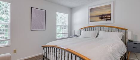 Un dormitorio con cama, dos mesitas de noche, una mesita auxiliar, una ventana y un cuadro en la pared.