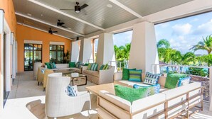 A covered outdoor lounge area with seating and tables, large windows, and a view of palm trees and a pool.