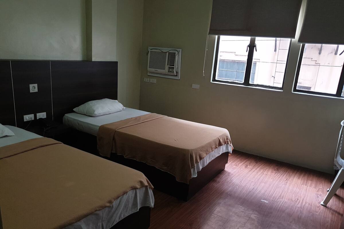 A hotel room with two single beds, a window with blinds, an air conditioning unit, and a wooden floor.
