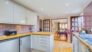 Een moderne keuken met witte kasten, een Kenwood koelkast en een houten aanrecht. Een eethoek met een tafel en stoelen is zichtbaar door een open deur.