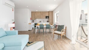 A modern apartment with a living room, dining area, and kitchen. It features a light blue sofa, a dining table with chairs, and a wicker chair by a large window.