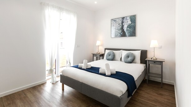 A modern bedroom with a large bed, two bedside tables with lamps, a window with sheer curtains, and a painting on the wall.