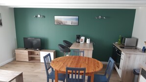 Ett kompakt vardagsrum med ett matbord i trä omgivet av stolar, en liten köksdel med mikrovågsugn och brödrost, en tv och en vägg som är målad i en accentfärg.