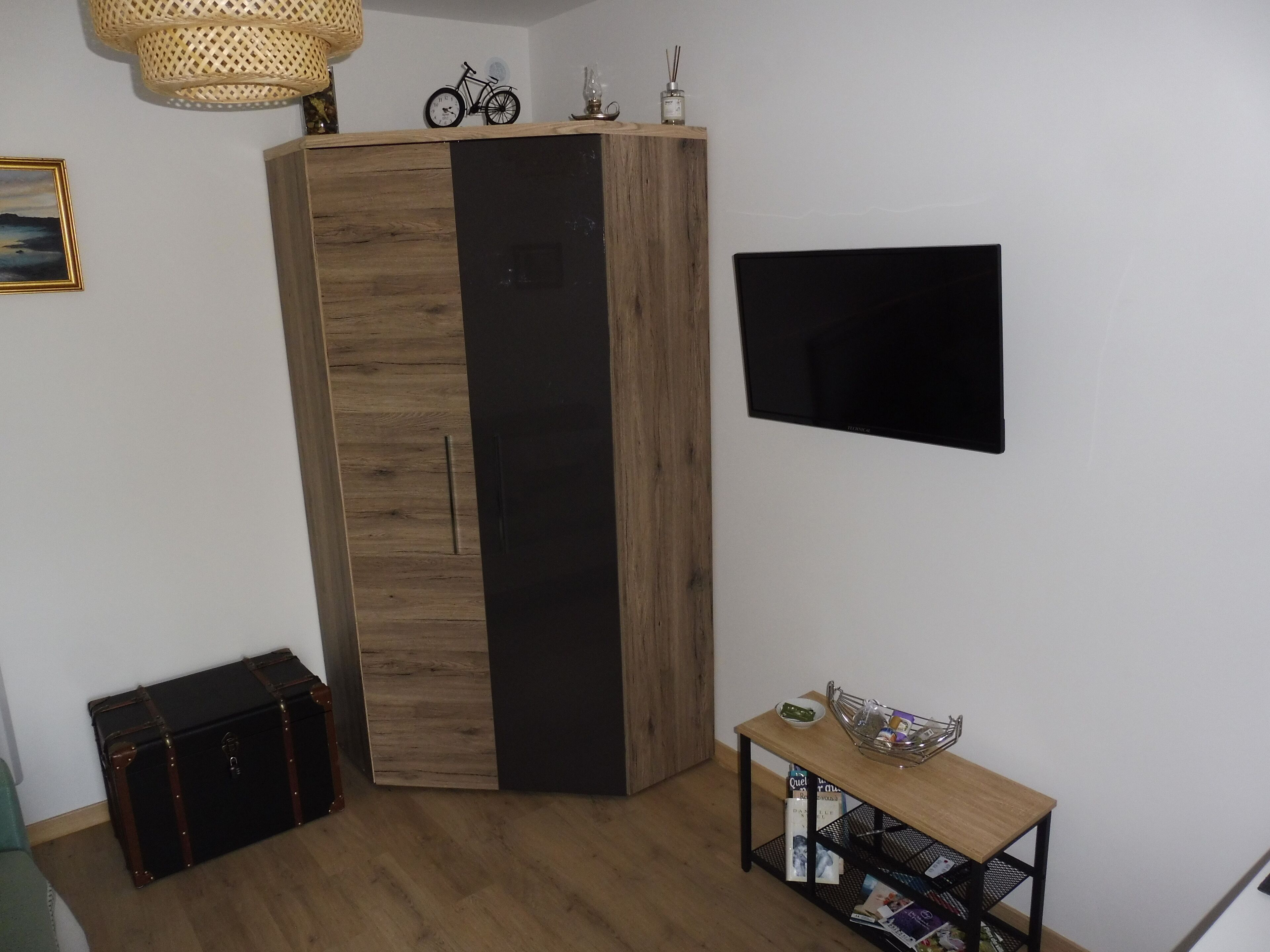 Una habitación con un armario de madera, un televisor negro en la pared, una pequeña mesa de madera con un bol y revistas, y una maleta en el suelo.