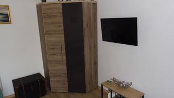 Una habitación con un armario de madera, un televisor negro en la pared, una pequeña mesa de madera con un bol y revistas, y una maleta en el suelo.