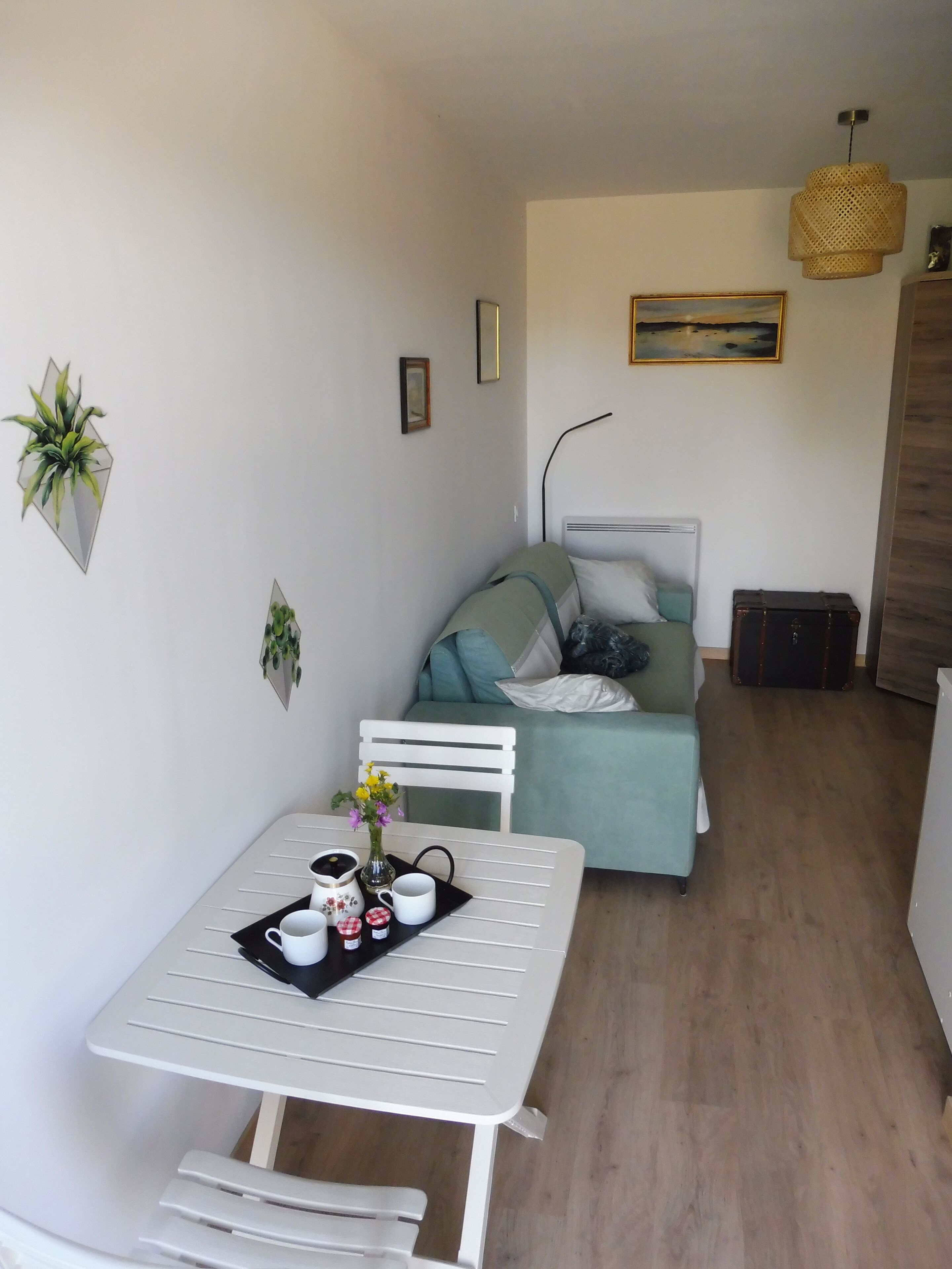 Un espace de vie compact comprenant une table à manger, un canapé et un lit. On y trouve également des plantes suspendues au mur et un tableau.