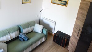Una sala de estar con un sofá verde, dos cuadros enmarcados, una lámpara de mimbre, un radiador y un mueble de madera.