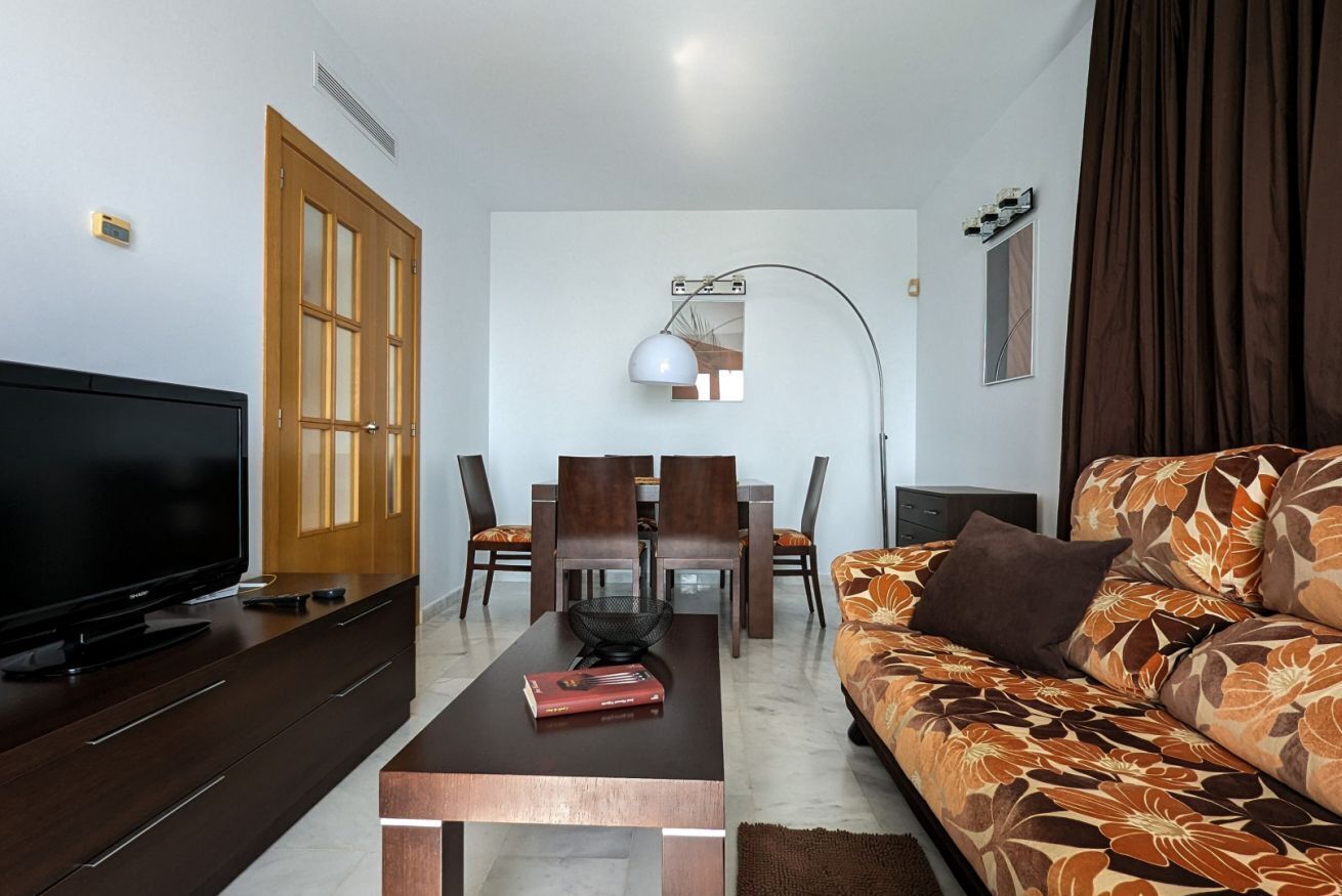 Een moderne woonkamer met een flatscreen-tv, een houten salontafel, een bank met een bloemenprint, een eethoek met een kroonluchter en een groot raam met gordijnen.