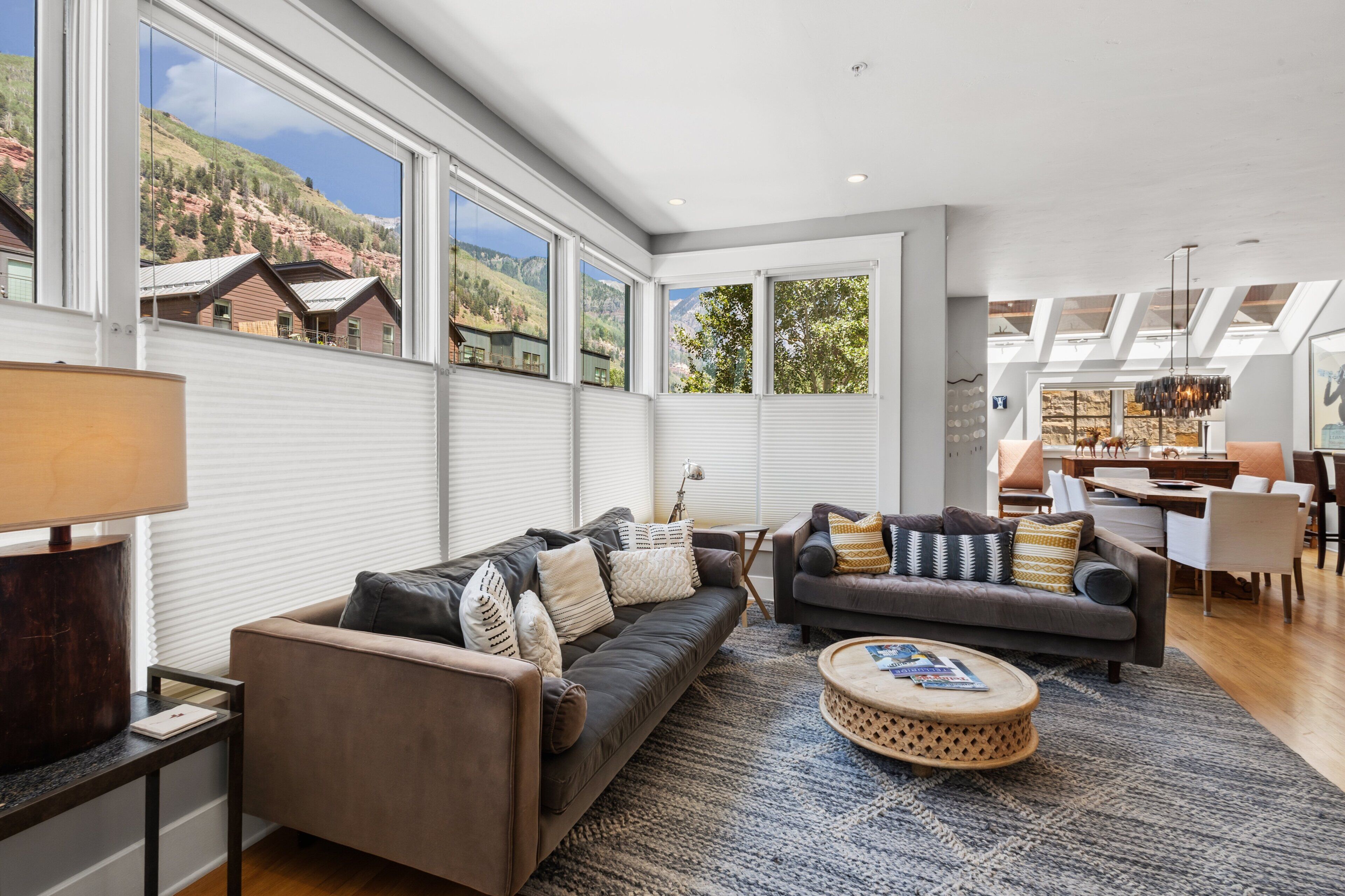 A spacious living room with a large window overlooking a mountainous landscape, a dark brown sofa, a grey sofa, and a round wooden coffee table.