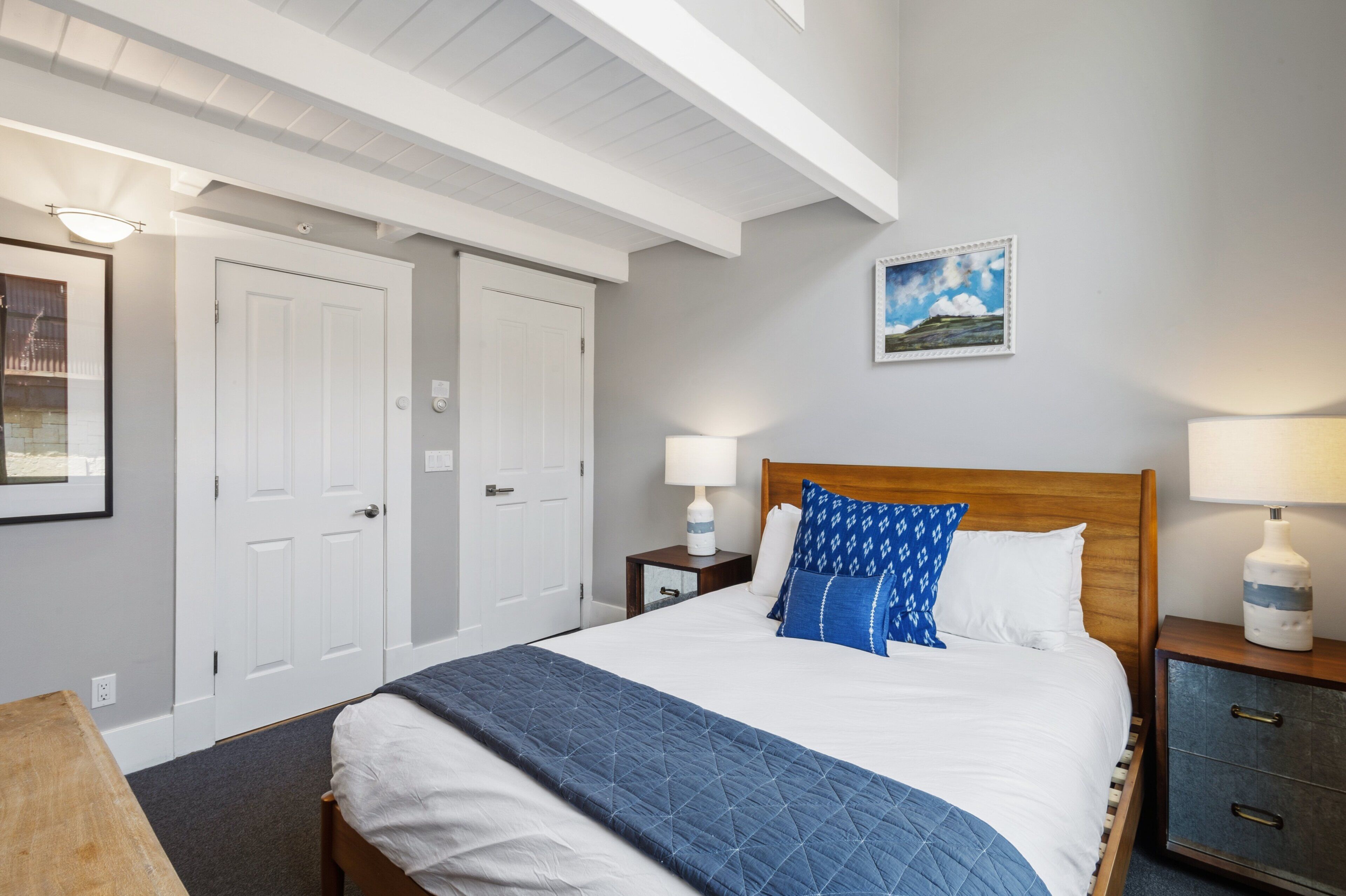 Ett hotellrum med en säng, två sängbord, ett litet bord, en träpanel vid sänggaveln, en tavla som hänger på väggen och en dörr.