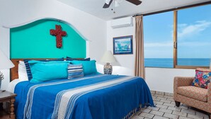 A bedroom with a bed, a cross on the headboard, a window with a view of the sea, and a chair with a colorful cushion. - Condominium Playa Bonita (Puerto Vallarta)