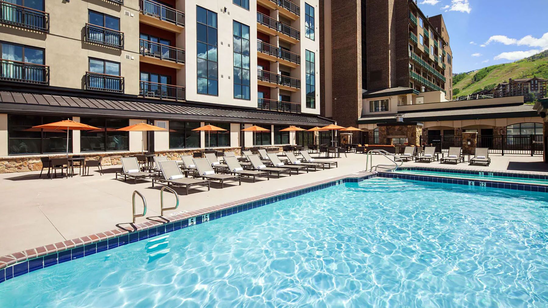 Un espace piscine d’hôtel avec des chaises longues et des parasols, une piscine à l’eau d’un bleu limpide et un bâtiment doté de nombreux balcons.