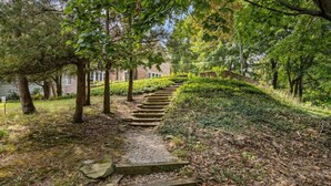 Eine steinerne Treppe führt einen grasbewachsenen Hang hinauf, umgeben von Bäumen und einem Gebäude im Hintergrund.