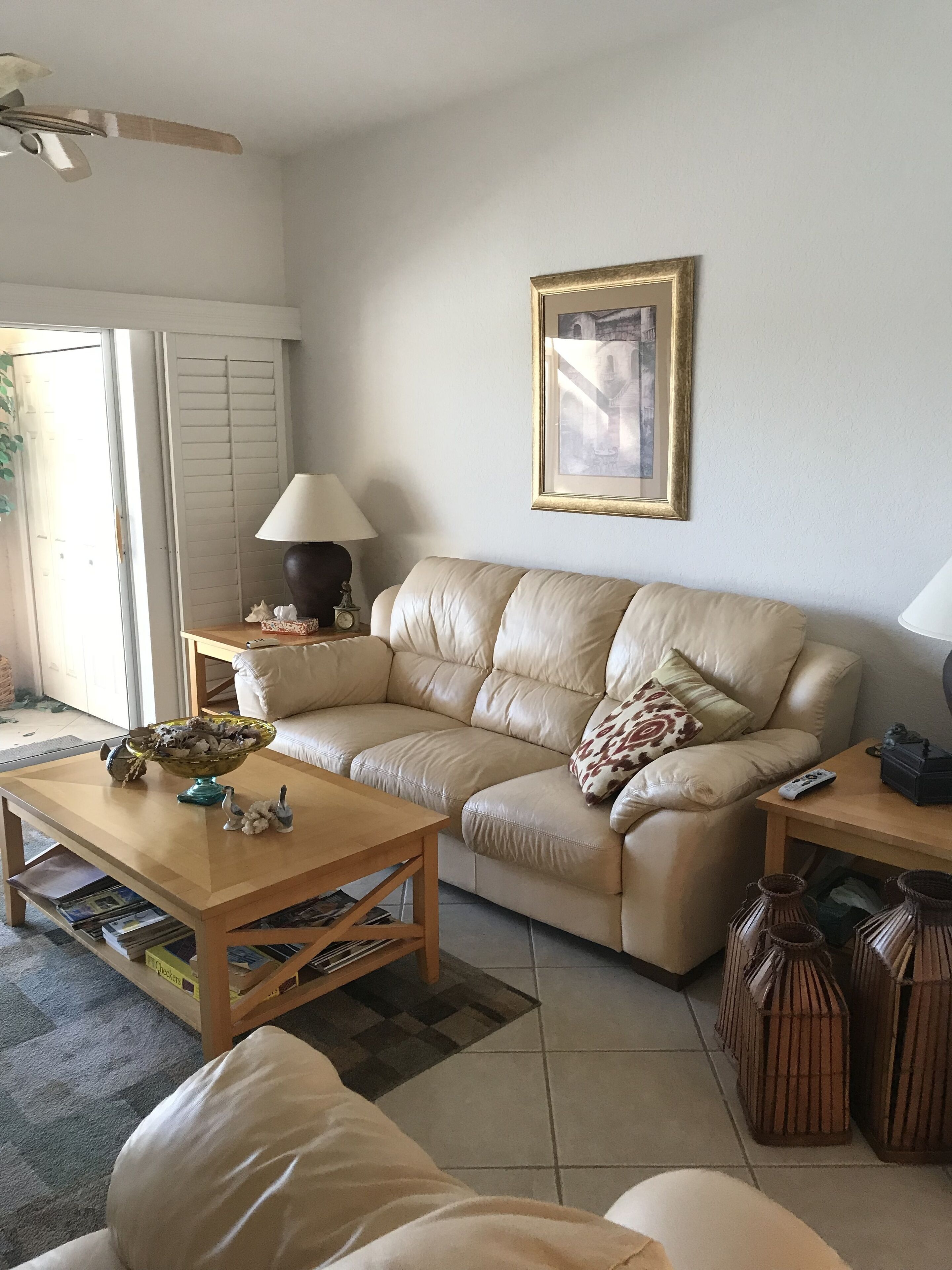 A living room with a beige leather sofa, a wooden coffee table, a lamp, a framed picture on the wall, and a bookshelf with magazines.