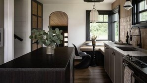 A modern kitchen with a central island, a dining area with a bench, and a view of the outdoors through large windows. - Hood Canal Beach Retreat (Belfair)