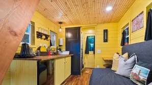 A compact kitchen with wooden cabinets, a sink, and a coffee maker. There is a small dining area with a table and a couch with pillows. - The Arthur - Custom Tiny Home w/ Train Vibe (Portland)