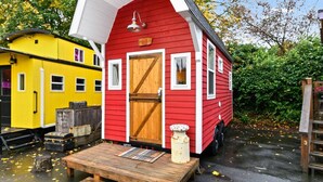Una casa móvil roja con puerta y ventanas de madera, un porche pequeño y un edificio amarillo al fondo.
