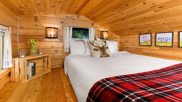A cozy wooden cabin bedroom with a bed, pillows, a nightstand, a window with curtains, and wall-mounted lamps.