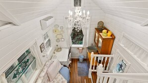 A narrow, white-paneled room with a chandelier, a small table with a candle, and a window with a view of greenery. - The Cottage - Custom Tiny Home w/ Victorian Vibe (Portland)