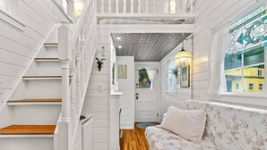 A room with a wooden staircase, a floral sofa, and a view of a yellow building through a window. - The Cottage - Custom Tiny Home w/ Victorian Vibe (Portland)