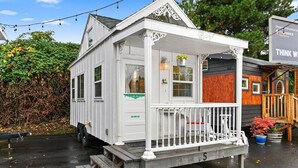 A white, single-story house with a porch, a small deck, and a sign indicating it is a cottage. - The Cottage - Custom Tiny Home w/ Victorian Vibe (Portland)