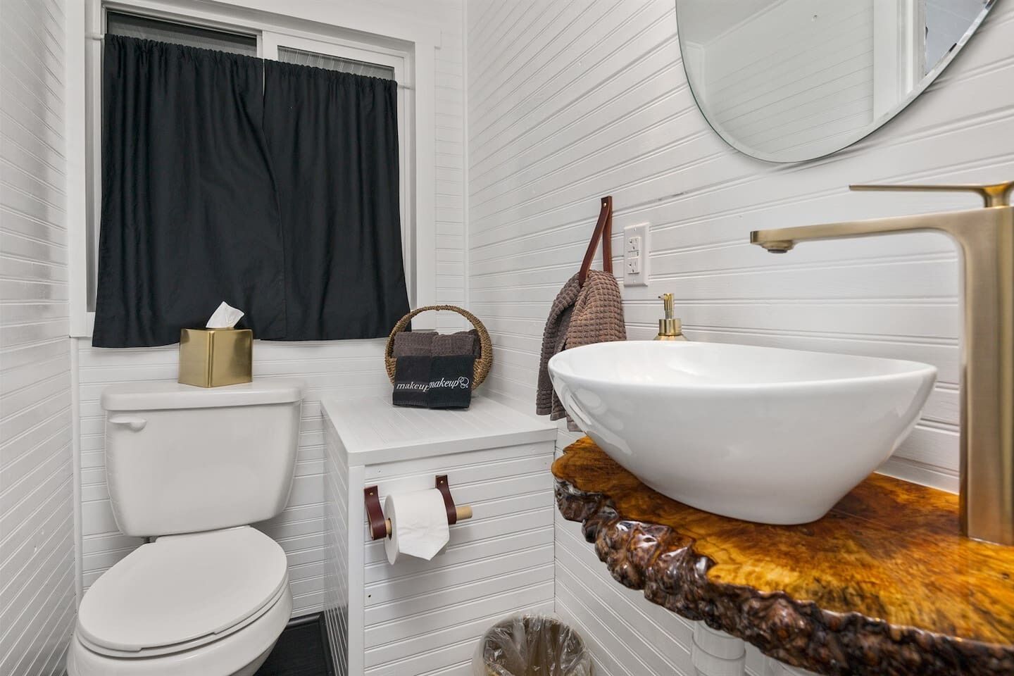 Un baño con inodoro blanco, un lavabo con un recipiente grande, un espejo sobre el lavabo y una ventana con cortinas negras.