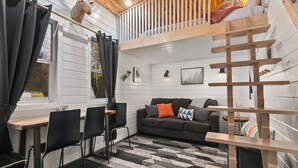 A cozy cabin interior with a loft bed, a dining area with chairs, a sofa with cushions, and a patterned rug. - The Finlandia - Custom Tiny Home w/ Finnish Vibe (Portland)