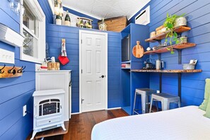 A compact room with blue walls, a white door, a small white stove, a bed with a white duvet, and shelves with various items.