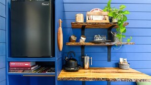 Una cocina con un refrigerador negro, estantes de madera con utensilios de cocina, una tetera y una pequeña mesa con una planta.