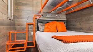A compact bedroom with a bed, orange pillows, a grey quilted bedspread, and a wooden floor. - The Modern - Custom Tiny Home w/ Modern Vibe (Portland)